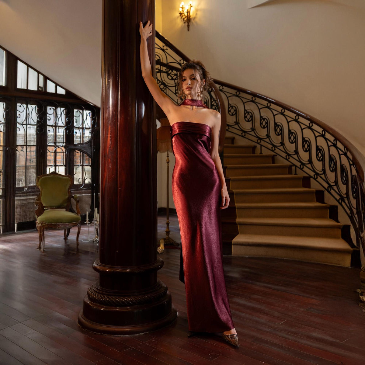 May include: A floor-length, strapless burgundy dress with a subtle sheen. The dress features a fitted silhouette and a high neckline. The image is set in a room with a wooden floor, a spiral staircase, and a large, dark wooden pillar.