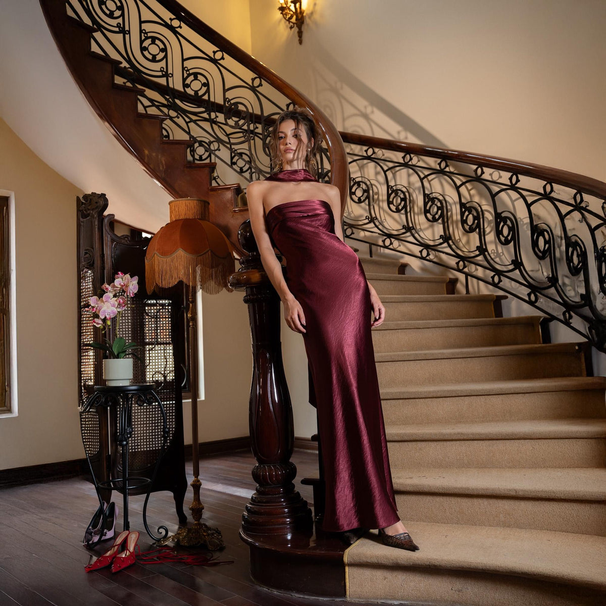 May include: A burgundy-colored, strapless, floor-length gown. The dress has a silky texture and a high neckline. The model is leaning against a dark wooden staircase with ornate wrought iron railings. The background includes a vintage lamp and a small table with flowers.