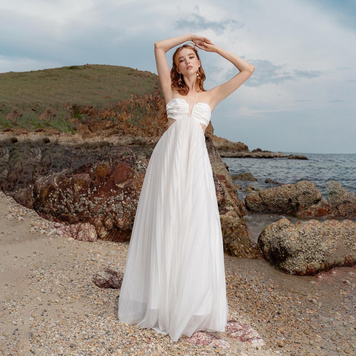 May include: A strapless white gown with a flowing skirt. The dress features cut-out details at the waist and a sweetheart neckline. The model is standing on a sandy beach with arms raised, with the ocean and rocks in the background.