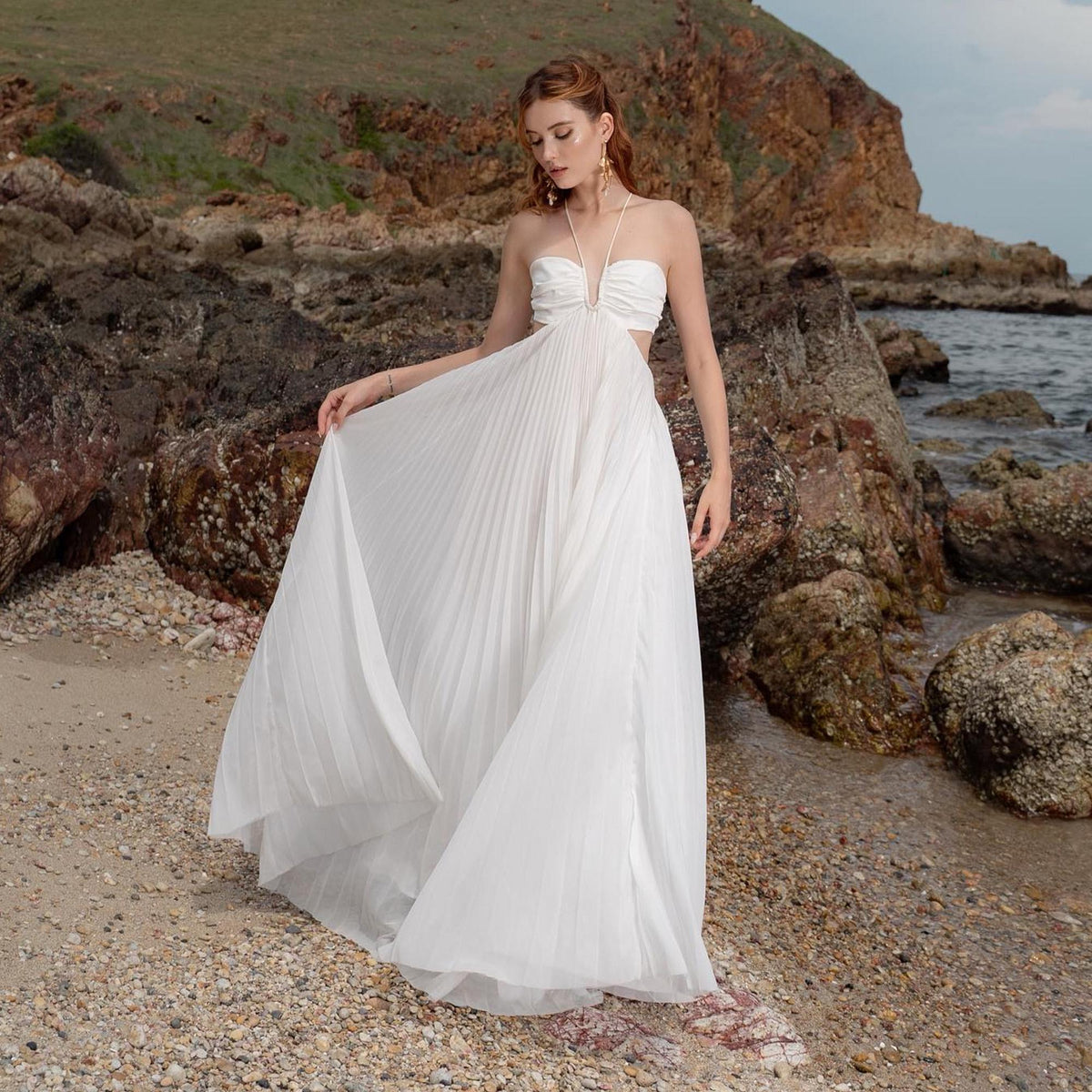 May include: A white pleated maxi dress with a halter neckline and cut-out details. The dress is flowing and elegant, perfect for a beach or outdoor event. The model is standing on a sandy beach with rocks and the ocean in the background.