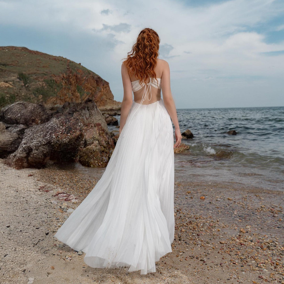 May include: A white, flowing gown with a halter top and open back, worn on a beach. The dress has a long, pleated skirt that trails behind the wearer. The person has long, red hair. The background includes the ocean, rocks, and a cloudy sky.