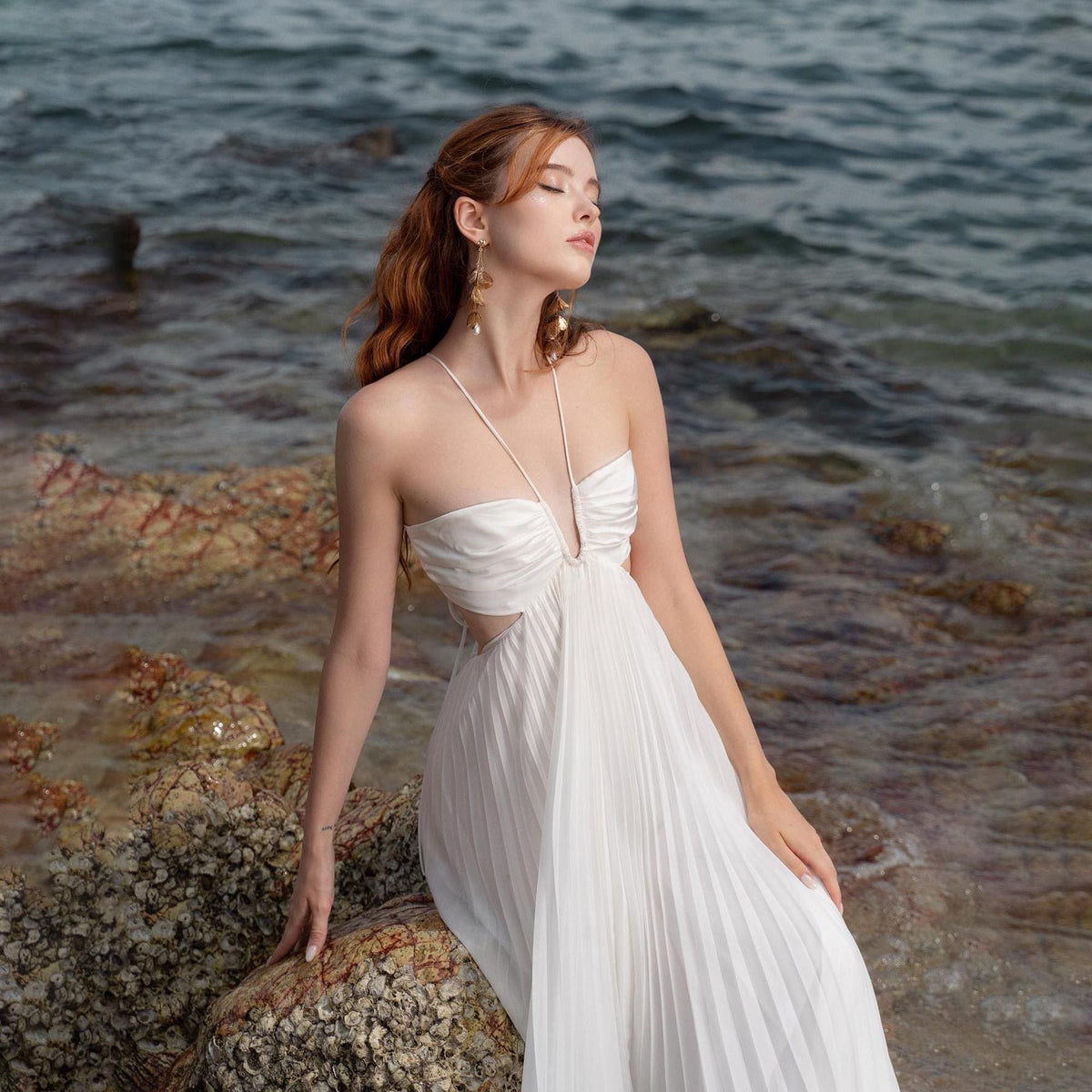 May include: A white, pleated maxi dress with a halter neckline and cut-out sides. The dress is worn by a person with auburn hair and gold earrings, posing on a rocky shoreline with the ocean in the background.