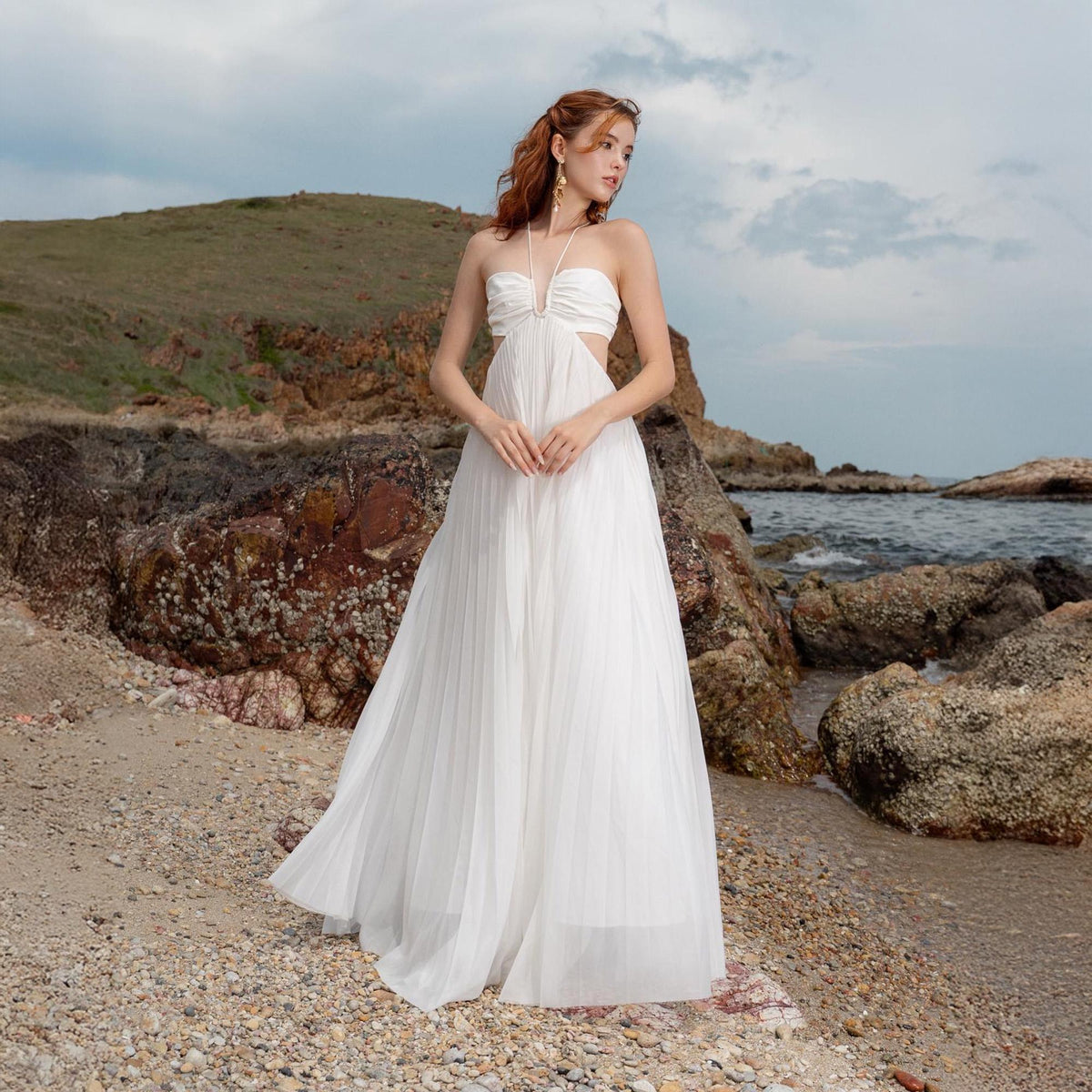 May include: A white halter-neck gown with a pleated skirt. The dress features cut-out details at the waist and a flowing, floor-length design. The model is standing on a beach with rocks and the ocean in the background.