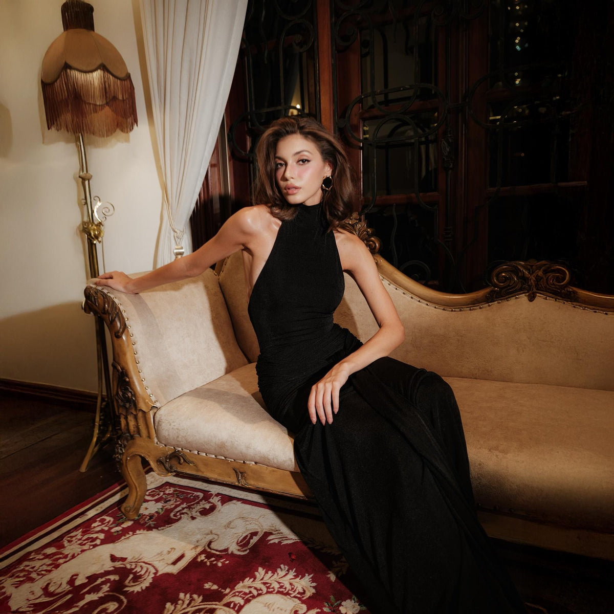 May include: A black halter-neck gown with a flowing silhouette. The dress features a high neckline and a sleeveless design. The model is seated on a vintage-style beige sofa with ornate detailing, set against a backdrop of a room with a warm, inviting ambiance.