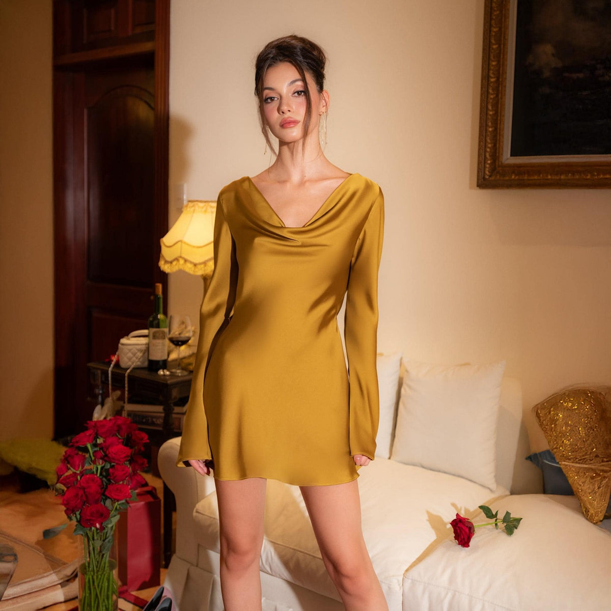 May include: Mustard yellow satin mini dress with long sleeves and a cowl neckline. The dress is a solid color and has a slight sheen. The model is standing in front of a white couch and a table with a lamp.