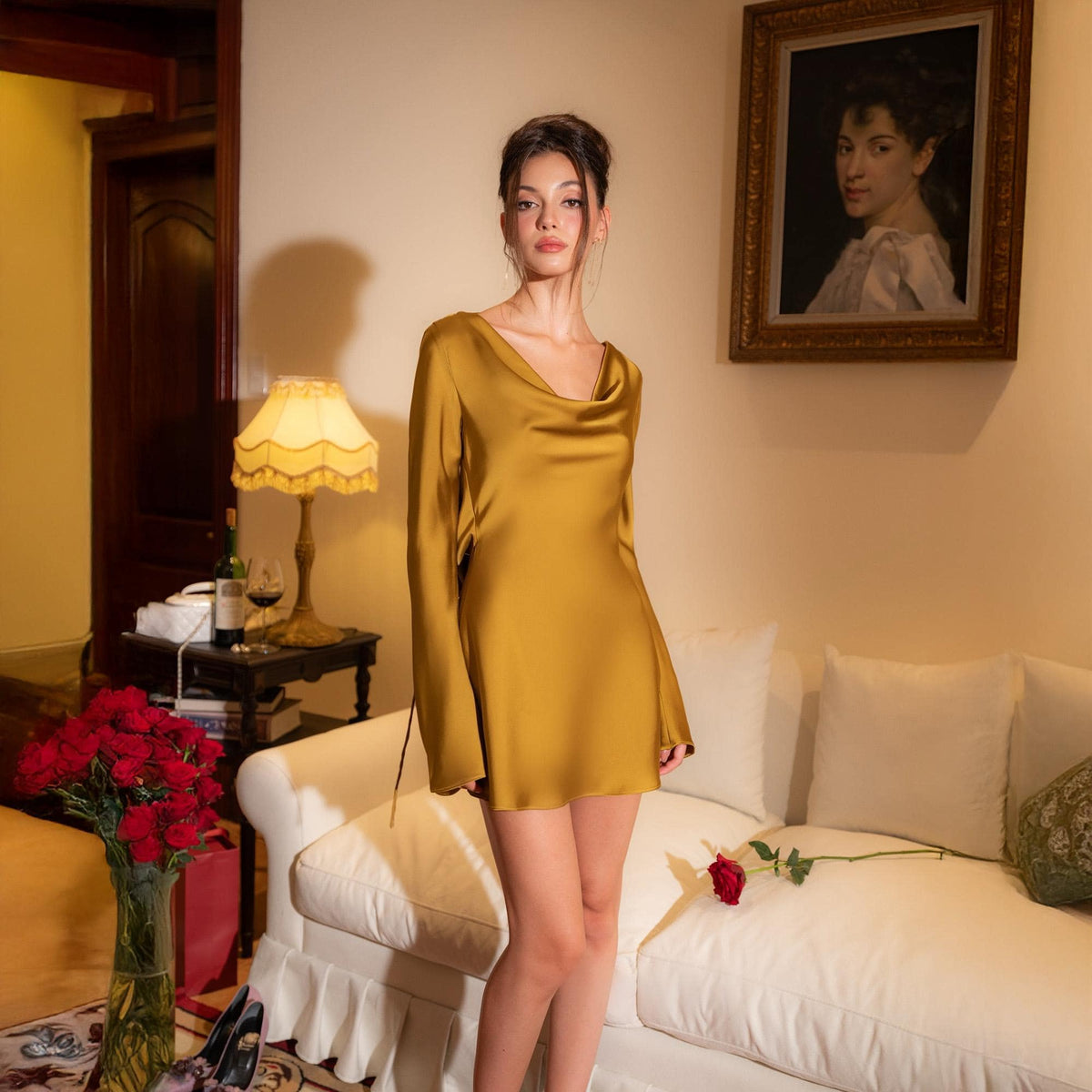 May include: A woman wearing a gold-colored satin mini dress with long sleeves and a cowl neckline. The dress is knee-length. The background includes a white sofa, a painting, a lamp, and a vase of red roses.