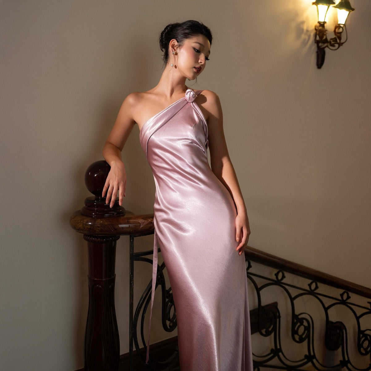 May include: A light pink, one-shoulder satin gown. The dress features a flowing, floor-length design with a subtle sheen. A decorative flower detail accents the shoulder. The model is leaning against a dark wooden banister.