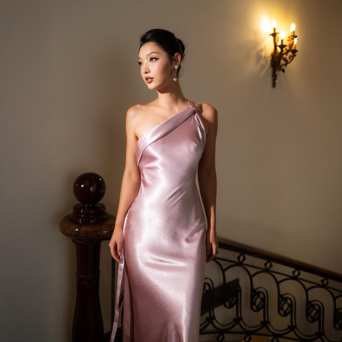May include: A light pink, one-shoulder satin gown. The dress has a flowing, floor-length design with a side slit. The model is standing on a staircase with a dark wooden banister and a decorative wall sconce.