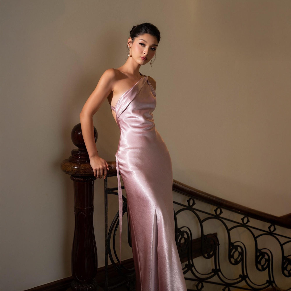 May include: A light pink, one-shoulder formal gown with a thigh-high slit. The dress has a flowing, satin-like texture and is worn by a person standing near a wooden banister. The background is a neutral color.
