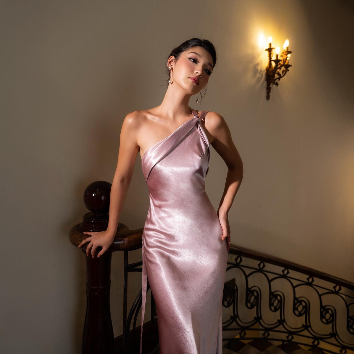 May include: A light pink, one-shoulder satin gown. The dress has a draped neckline and a flowing silhouette. The model is standing on a staircase with a dark wooden banister. A wall sconce with three lights is visible in the background.