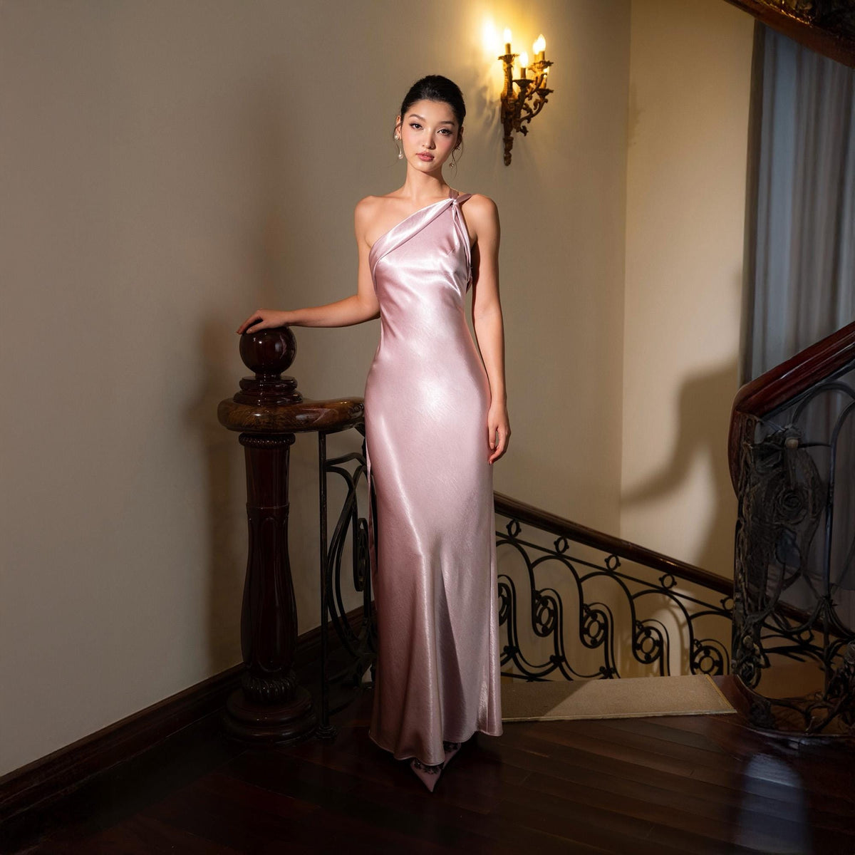 May include: A pale pink, one-shoulder satin gown. The dress has a flowing, floor-length design. The model is standing on a wooden staircase with a dark brown handrail. A decorative wall sconce with three lights is visible in the background.