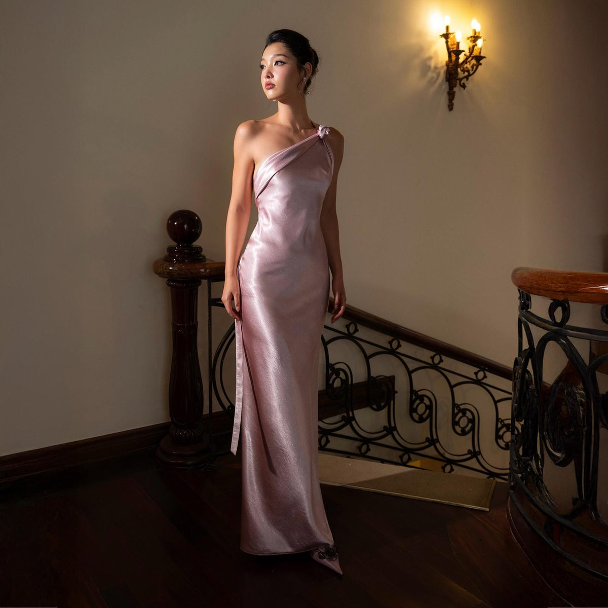 May include: A light pink, one-shoulder formal gown. The floor-length dress has a draped bodice and a flowing skirt. The dress is made of a shiny, silky material. The background includes a staircase with a dark wood banister and a wall sconce.