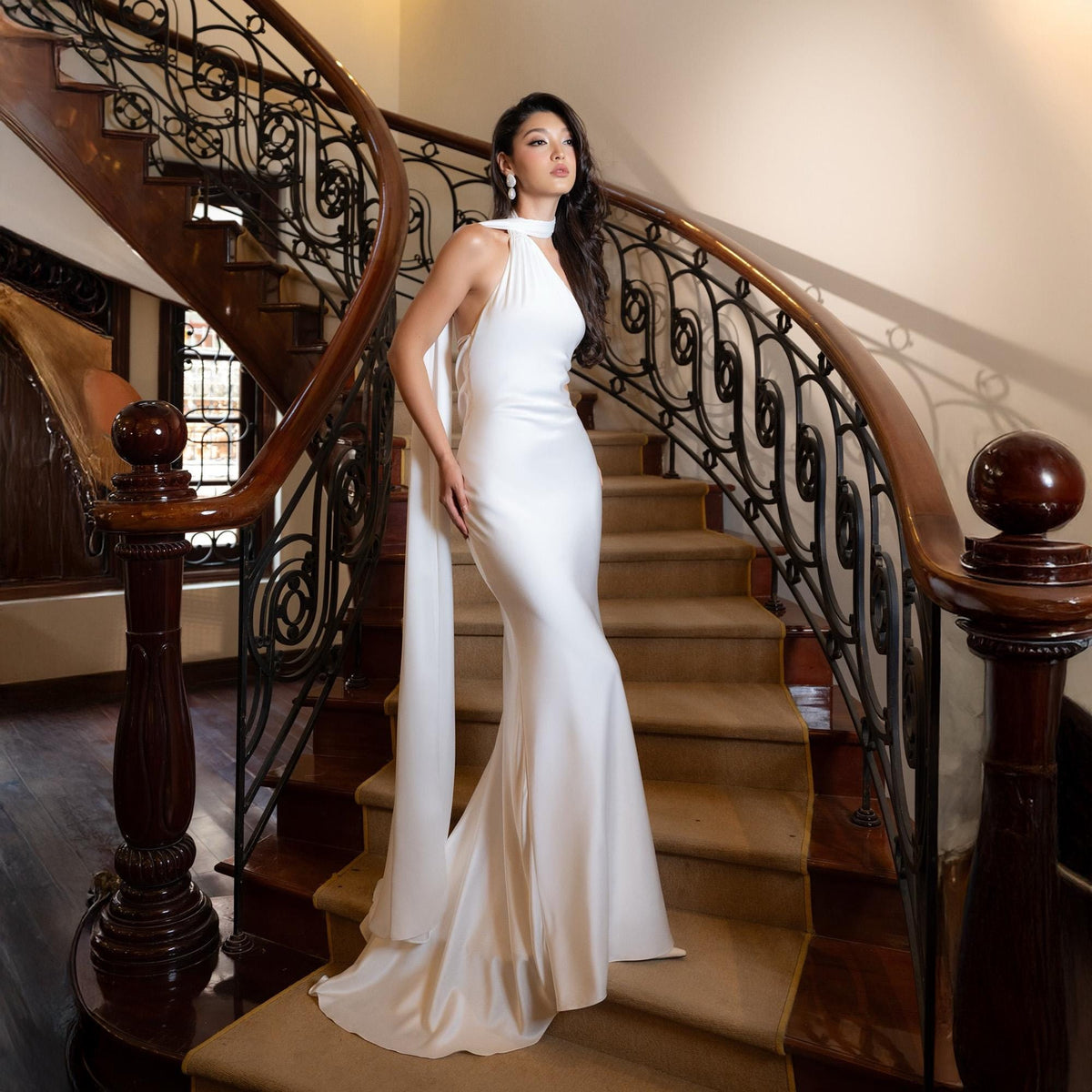 May include: A white, floor-length gown with a halter neckline and flowing cape detail. The dress is made of a smooth, silky fabric and is worn on a staircase with a dark wood banister and ornate metal railing. The overall aesthetic is elegant and formal.