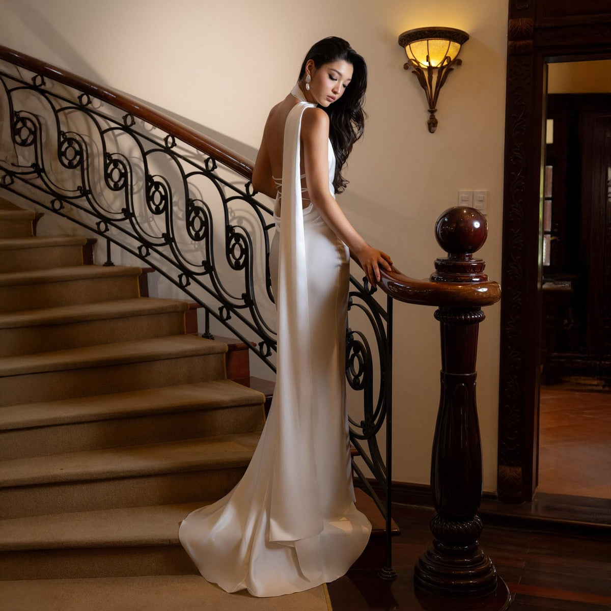 May include: A white, floor-length gown with a flowing train and a halter neckline. The dress features a draped back and is worn on a staircase with a decorative wrought iron railing. The setting is a well-lit interior with a classic design.