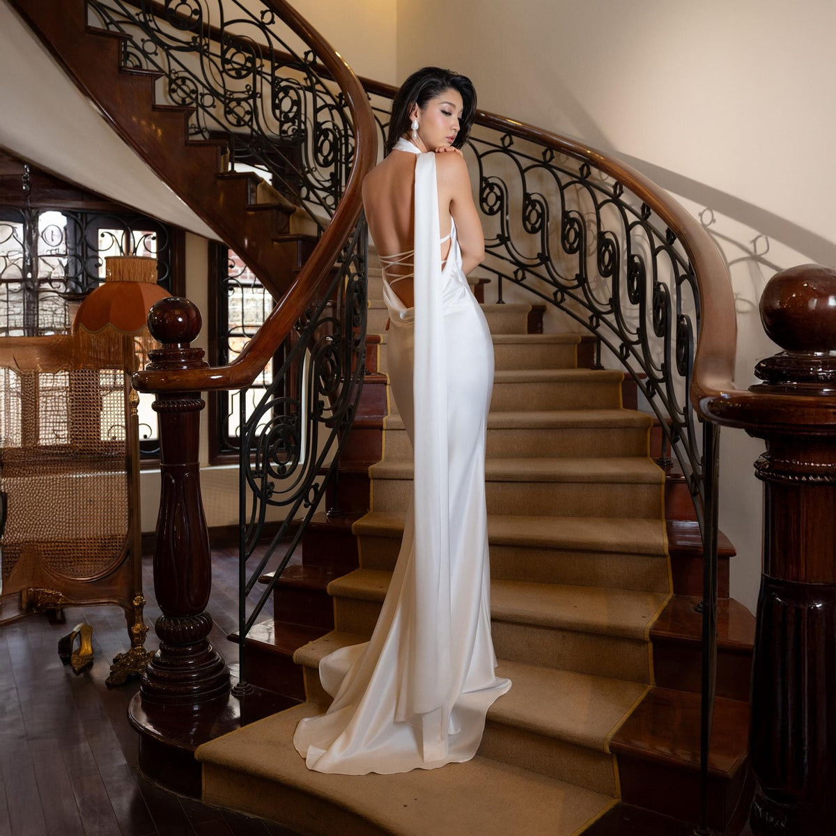 May include: A white, floor-length gown with a halter neckline and open back, featuring a flowing cape detail. The dress is worn on a staircase with a dark wood banister and ornate ironwork. The overall aesthetic is elegant and formal.