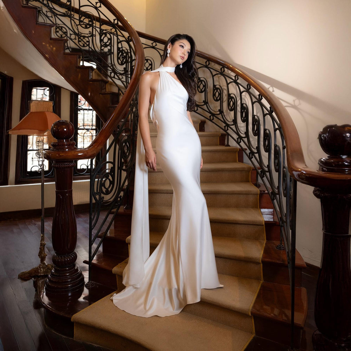 May include: A white, floor-length gown with a halter neckline and flowing train. The dress is worn on a staircase with a dark wooden banister and ornate metalwork. The stairs are covered in a neutral-toned carpet.