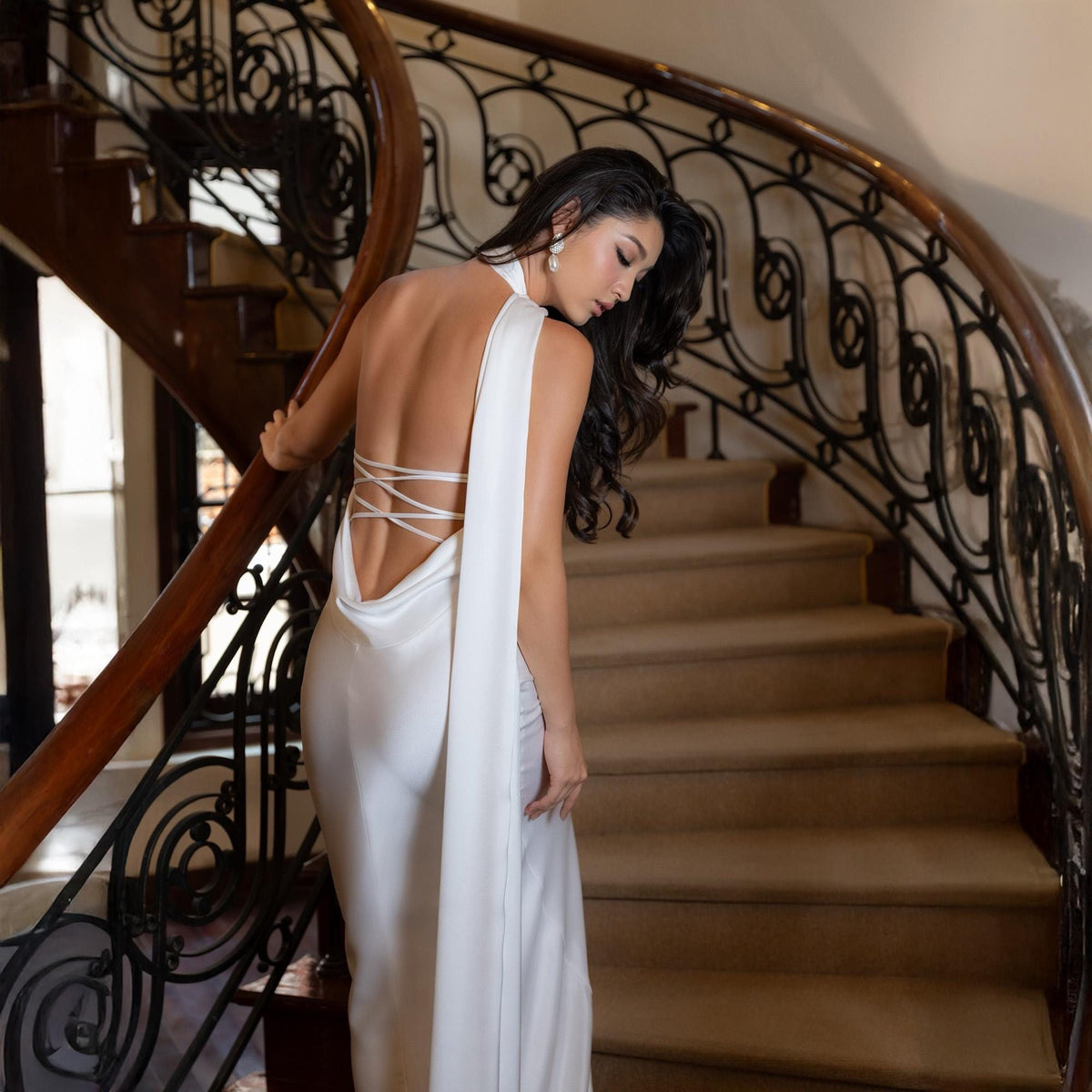 May include: A white satin gown with a halter neckline and a low, open back. The dress features a flowing, draped design and a lace-up back detail. The model is standing on a curved staircase with a dark wooden handrail and ornate metal balustrade.