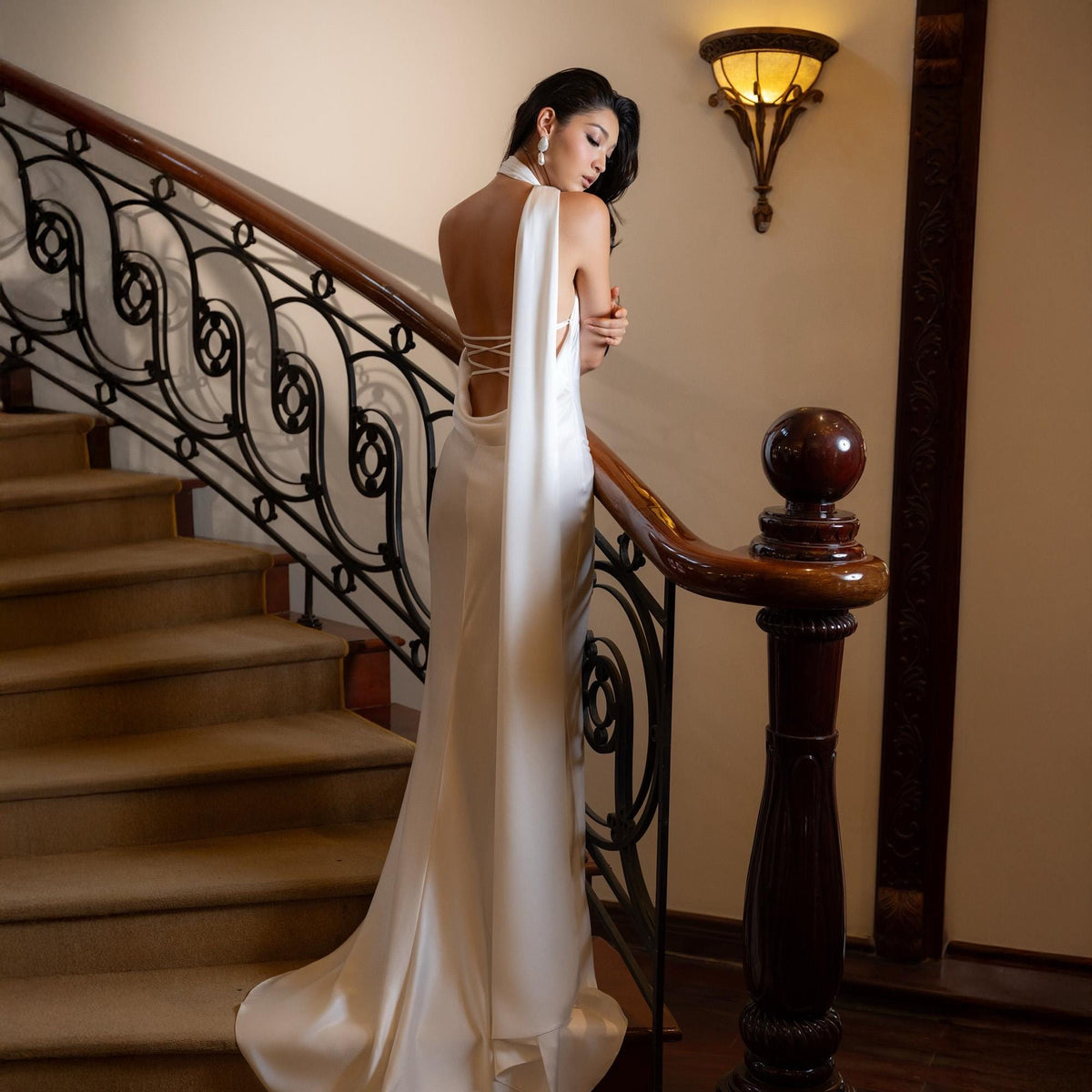 May include: A white satin gown with a draped back and a flowing train. The dress features a halter neckline and a low back with crisscross detailing. The model is standing on a staircase with a dark wood banister.