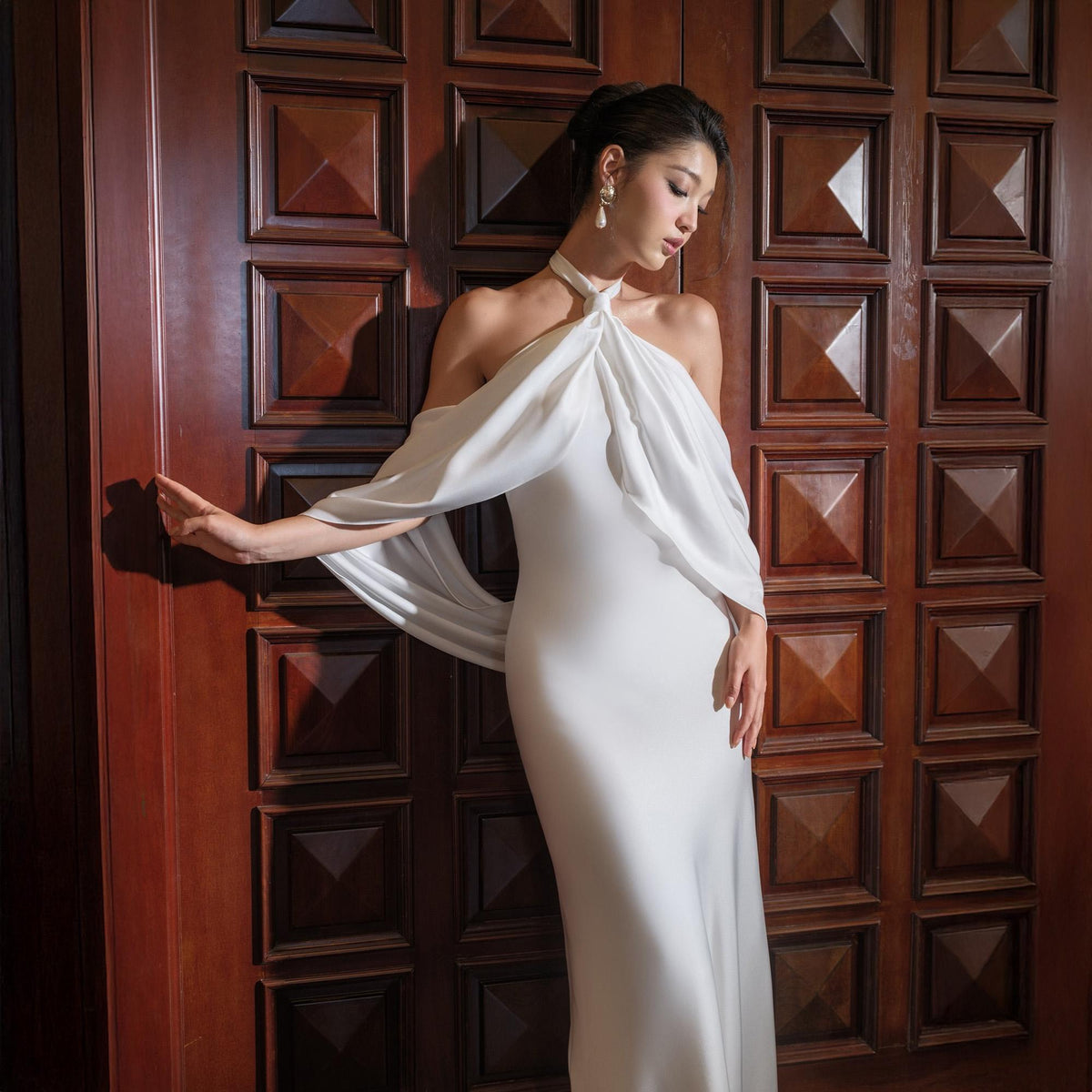 May include: A white, floor-length gown with a halter neckline and draped sleeves. The dress is form-fitting and made of a smooth fabric. The model is leaning against a dark wooden door.