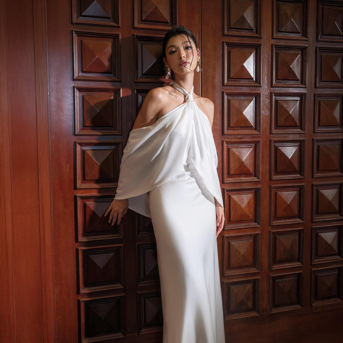 May include: A white, floor-length gown with a halter neckline and off-the-shoulder sleeves. The dress is made of a flowing fabric and has a simple, elegant design. The model is standing in front of a dark wooden door.
