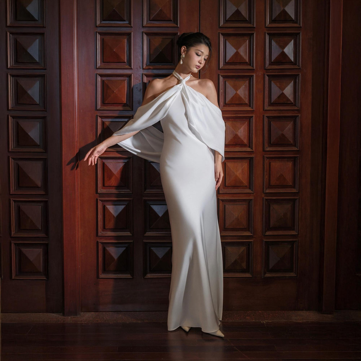 May include: A white, floor-length gown with a halter neckline and off-the-shoulder cape sleeves. The dress is made of a flowing fabric and is worn in front of a dark wooden door.