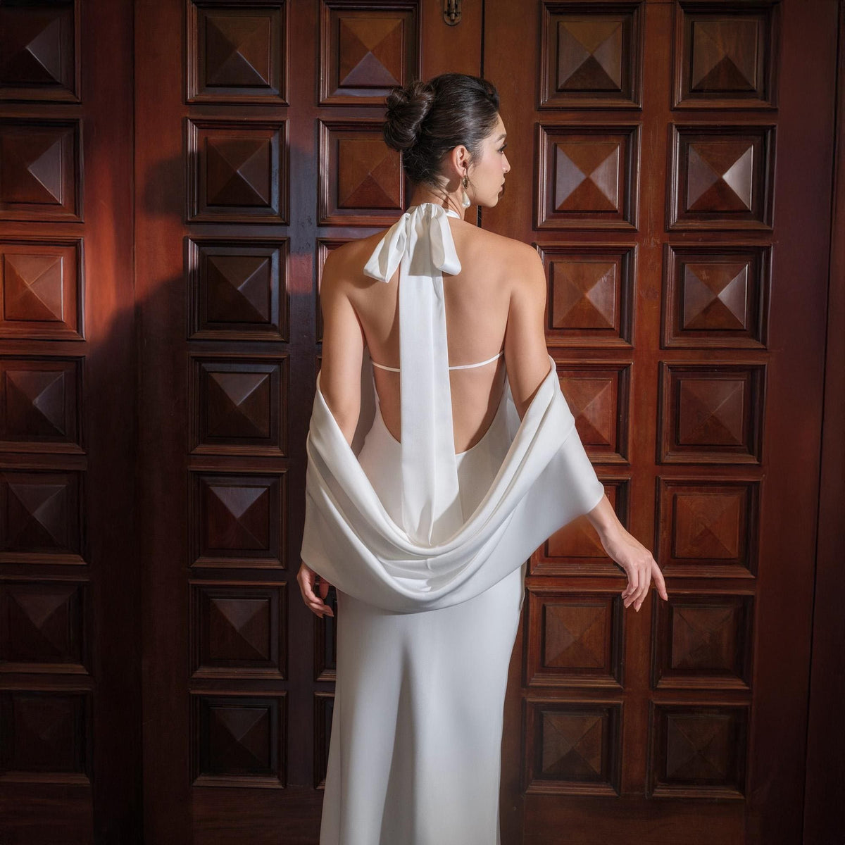 May include: A white halter-neck gown with a flowing cape. The dress has a low back and a long, elegant skirt. The model is standing in front of a dark wooden door.