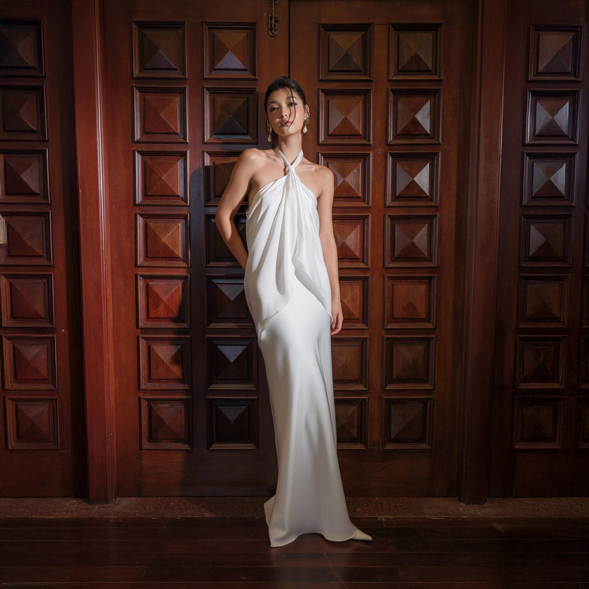 May include: A floor-length, white halter dress with a flowing silhouette. The dress features a draped bodice and a simple, elegant design. The model is standing in front of a dark wooden door.