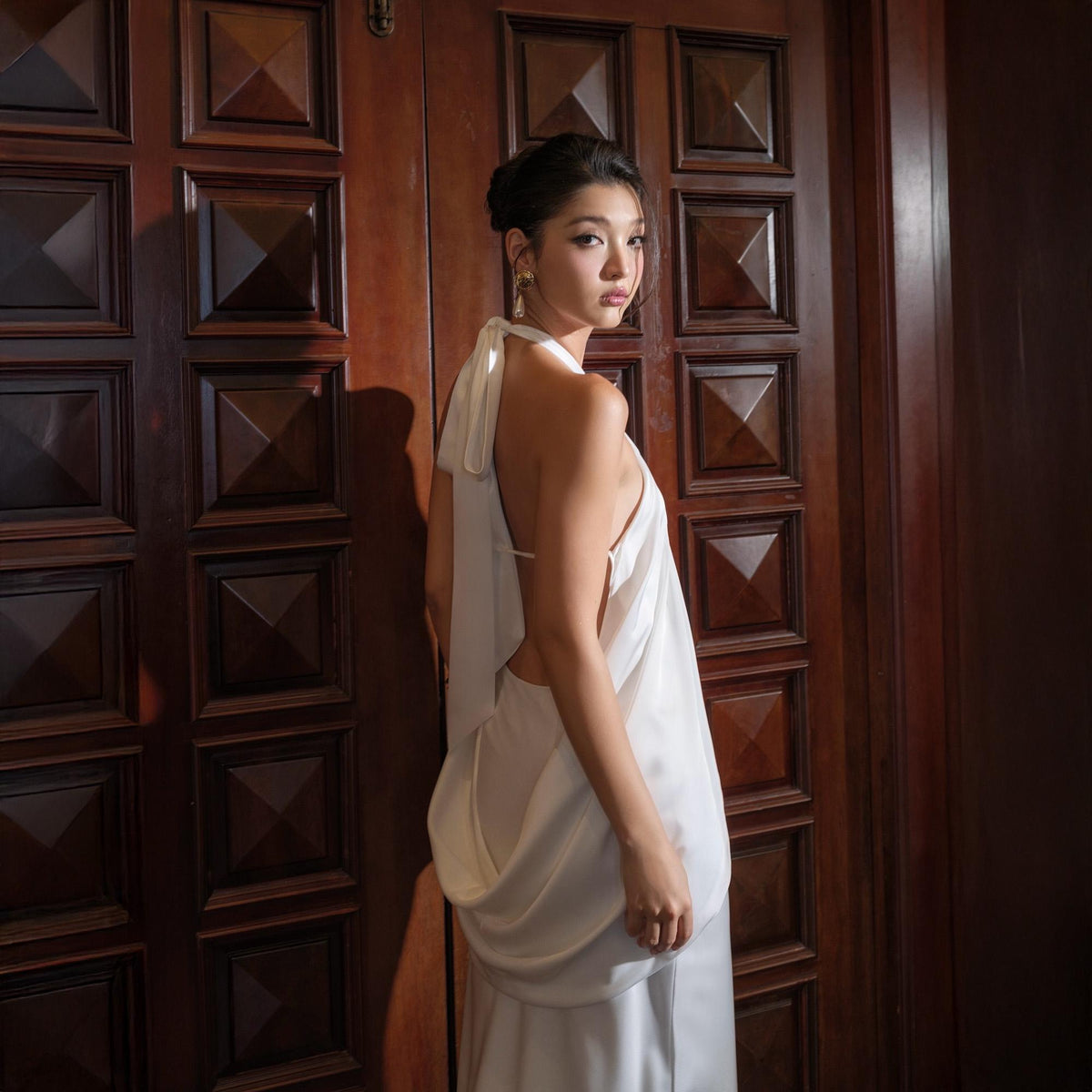 May include: A white, flowing halter dress with a draped cowl neckline and open back. The dress is worn by a person standing in front of a dark wooden door with square paneling. The dress has a simple, elegant design.