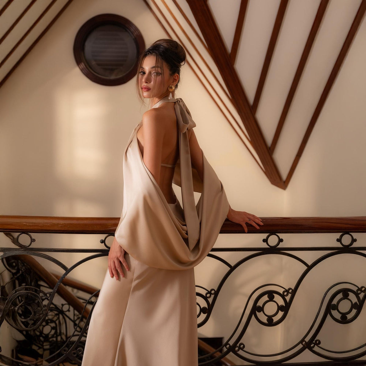 May include: A beige satin gown with a draped cowl neckline and open back. The dress features a flowing skirt and is accessorized with a bow at the neck. The model is standing near a decorative railing.