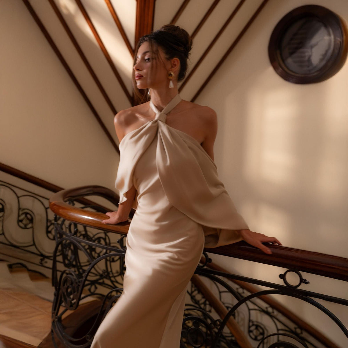 May include: A beige halter-neck gown with a flowing, draped design. The dress features a fitted silhouette and a soft, silky texture. The model is standing on a staircase with a wooden handrail and ornate metalwork.