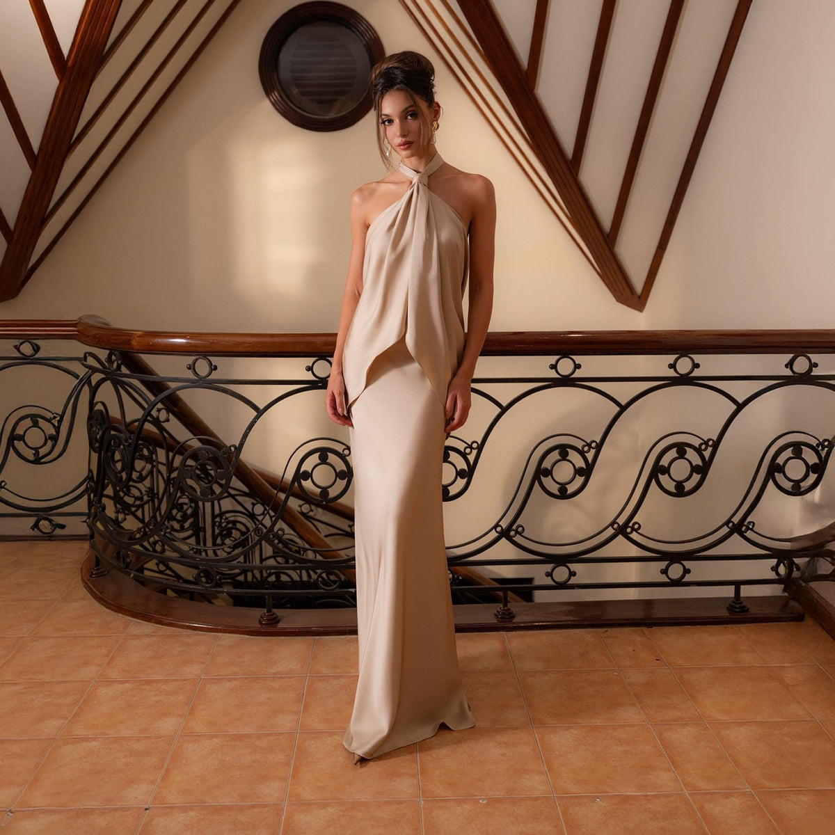 May include: A floor-length, halter-neck gown in a soft, neutral tone. The dress features a flowing skirt and a draped bodice. The model is standing on a tiled floor with a decorative wrought iron railing and wooden accents in the background.