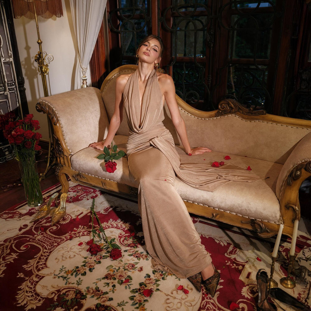 May include: A beige, floor-length gown with a draped cowl neckline. The dress is worn by a person seated on a beige chaise lounge. Red roses are scattered on the floor and a vase of roses is visible.