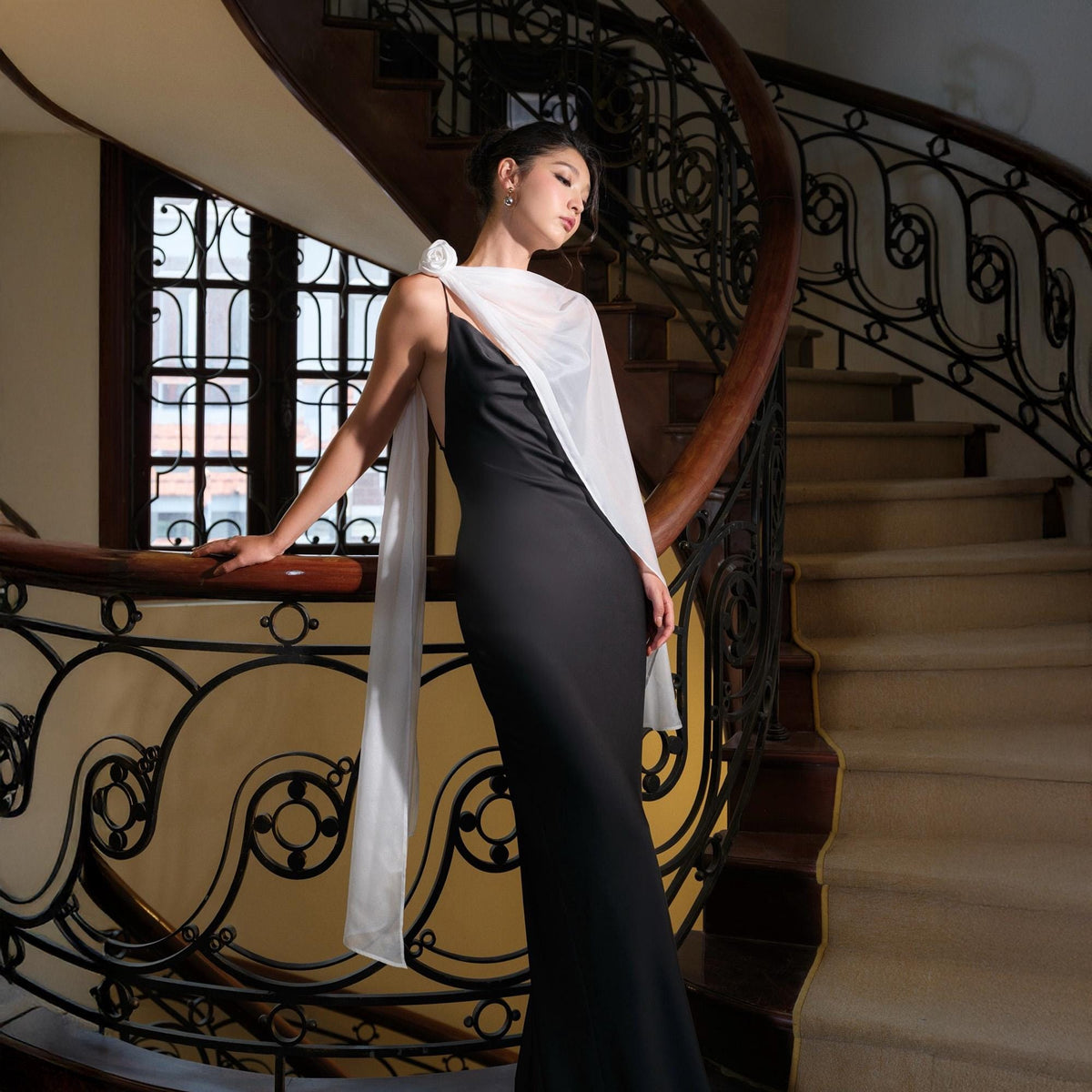 May include: A black, floor-length gown with spaghetti straps and a white scarf draped over the shoulder. The scarf is adorned with a white flower. The woman is standing on a wooden staircase with ornate wrought iron railings.