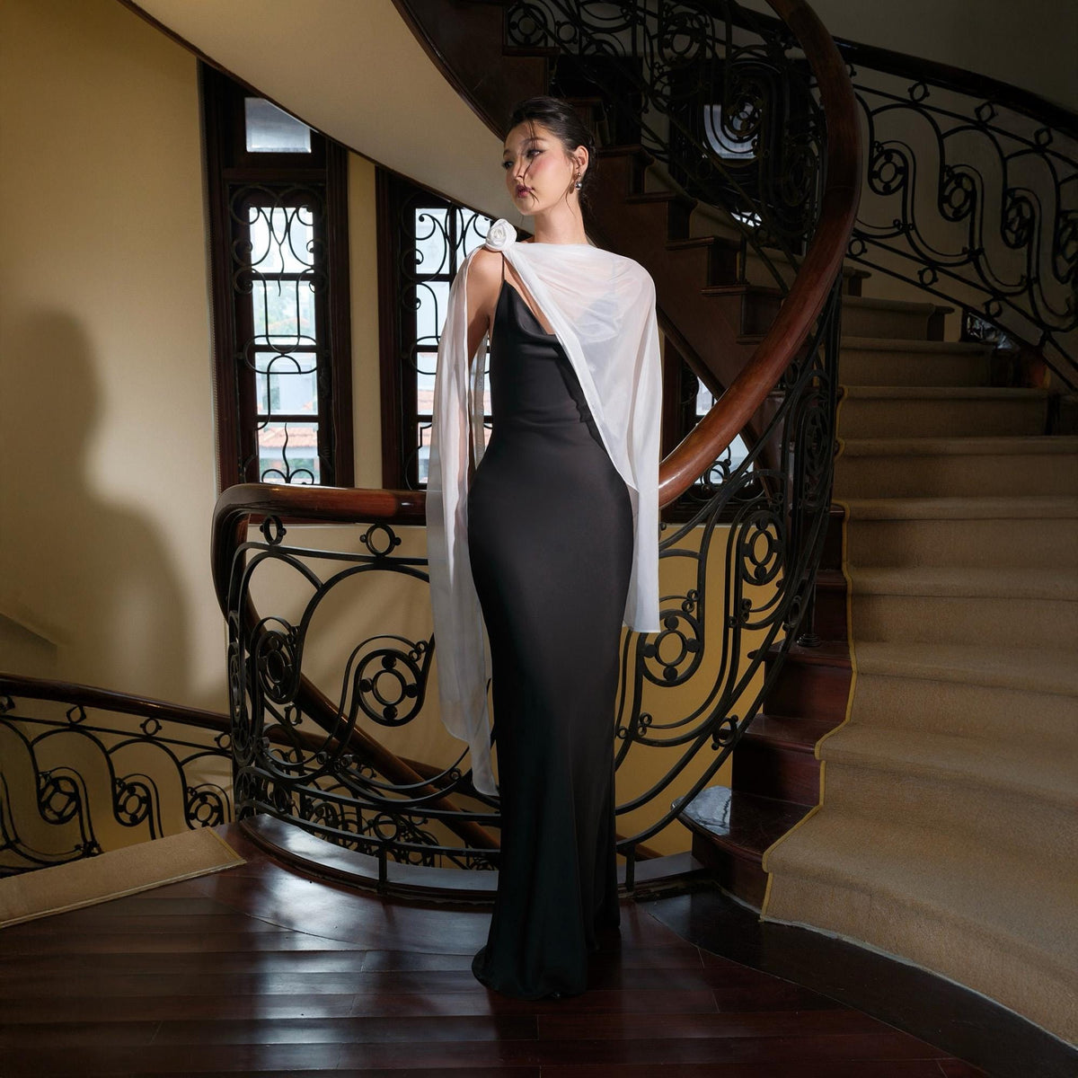 May include: A black, floor-length gown with a white, sheer shawl draped over the shoulders. The dress has a simple, elegant design. The background features a wooden staircase with ornate railings.