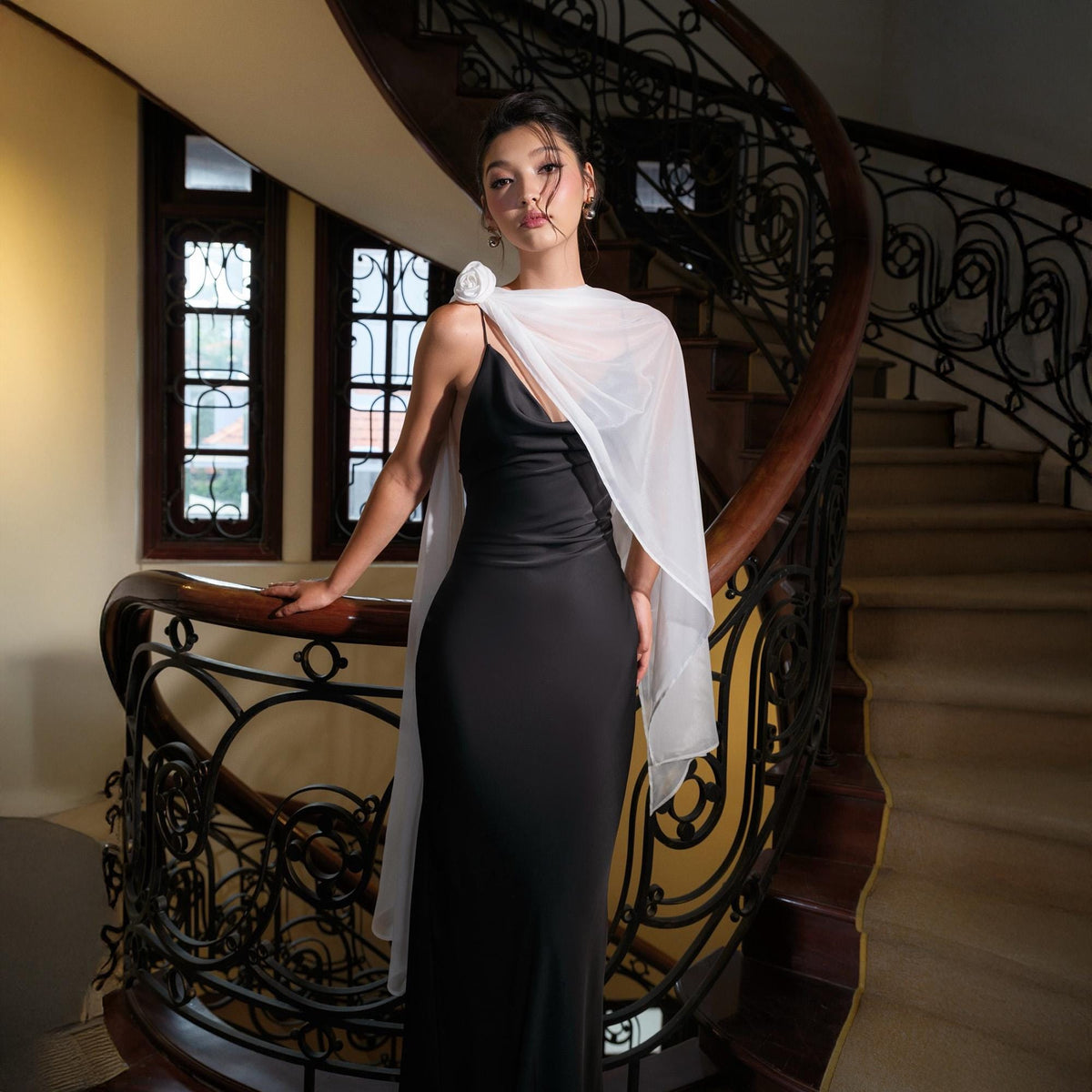 May include: A woman in a black, floor-length evening gown with spaghetti straps and a white, sheer shawl. A white flower adorns the shoulder. The woman is standing on a curved staircase with a dark wooden handrail and ornate metalwork.