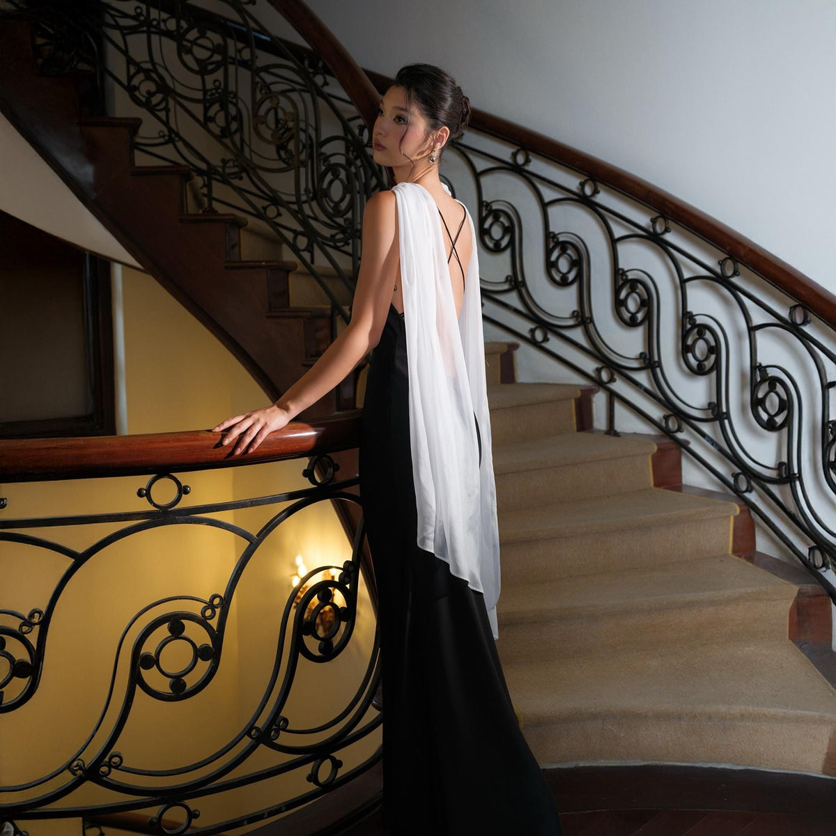 May include: A black formal gown with a white, flowing cape. The dress has a high slit and a low back. The model is standing on a staircase with a decorative wrought iron railing and wooden handrail.