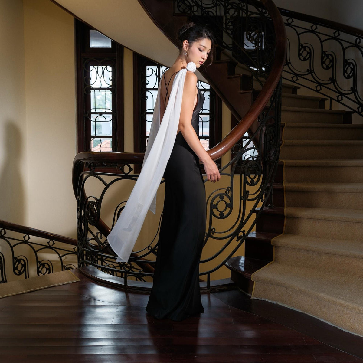 May include: A black jumpsuit with a white scarf-like overlay, accented with a white floral detail on the shoulder. The outfit is worn on a staircase with a dark wooden banister and light-colored walls.