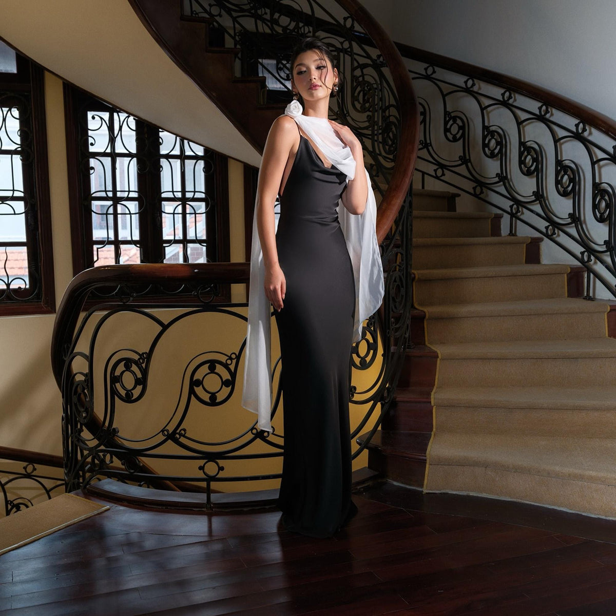 May include: A black, floor-length gown with spaghetti straps and a cowl neckline. A white, sheer scarf is draped over the shoulders. The woman is standing on a dark wood floor in front of a staircase with a decorative wrought iron railing.