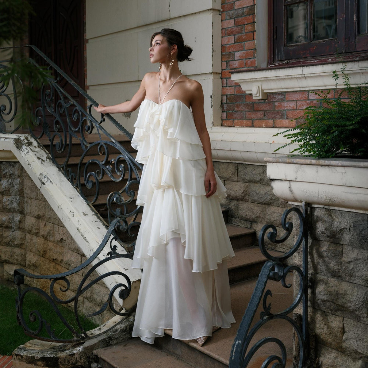 May include: A strapless, ivory-colored gown with layered ruffles. The dress features a flowing skirt and is worn outdoors. The model is standing on a stone staircase with a wrought iron railing.