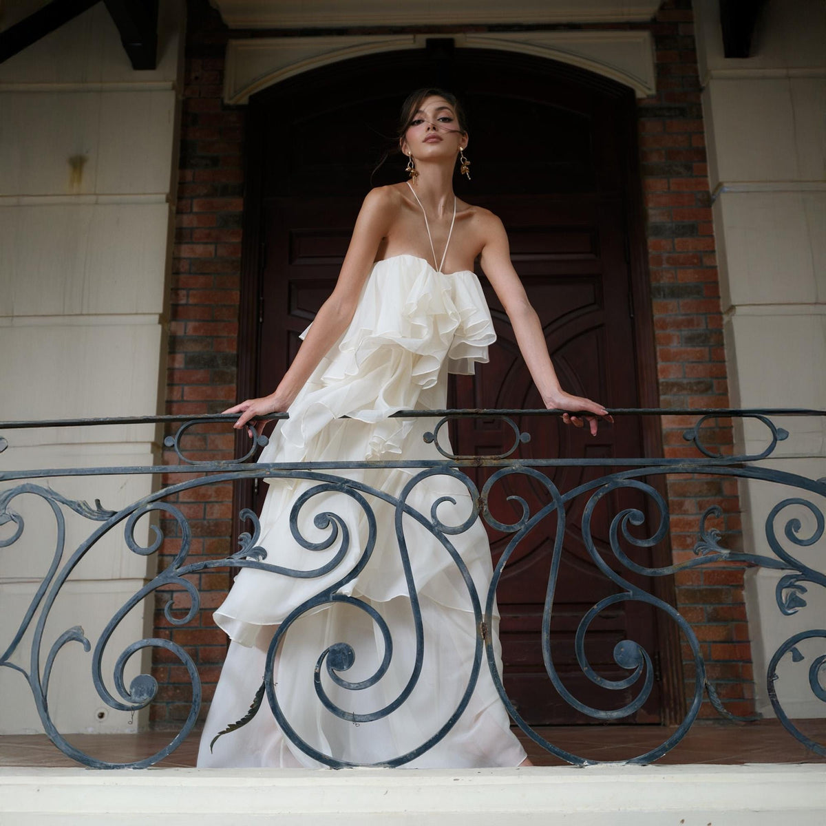 May include: A strapless ivory gown with layered ruffles and a flowing skirt. The dress is worn on a balcony with a decorative wrought iron railing. The background includes a brick wall and a dark wooden door.