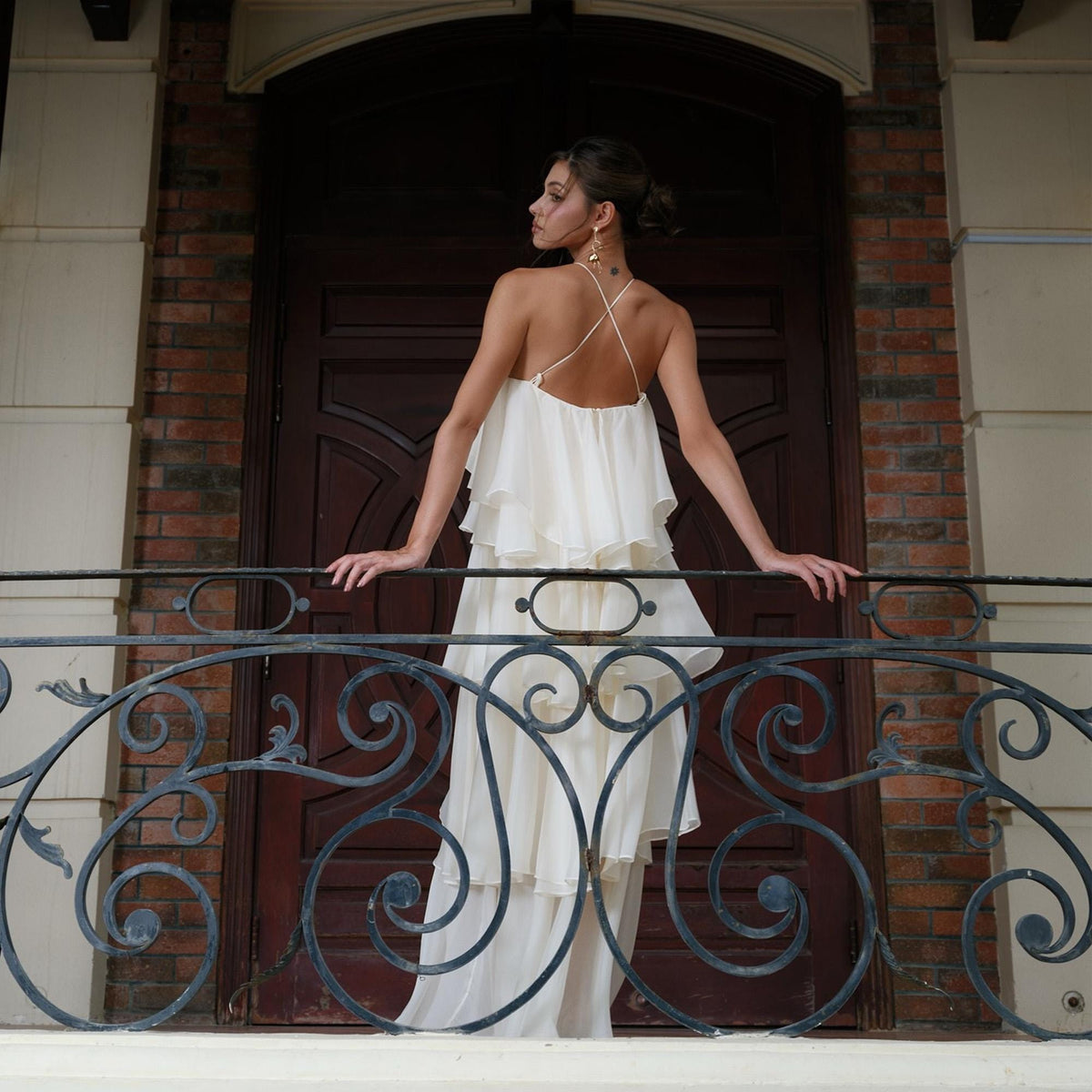 May include: A cream-colored, tiered maxi dress with spaghetti straps and a flowing skirt. The dress is worn on a balcony with a decorative wrought iron railing. The background features a dark wooden door and brick wall.