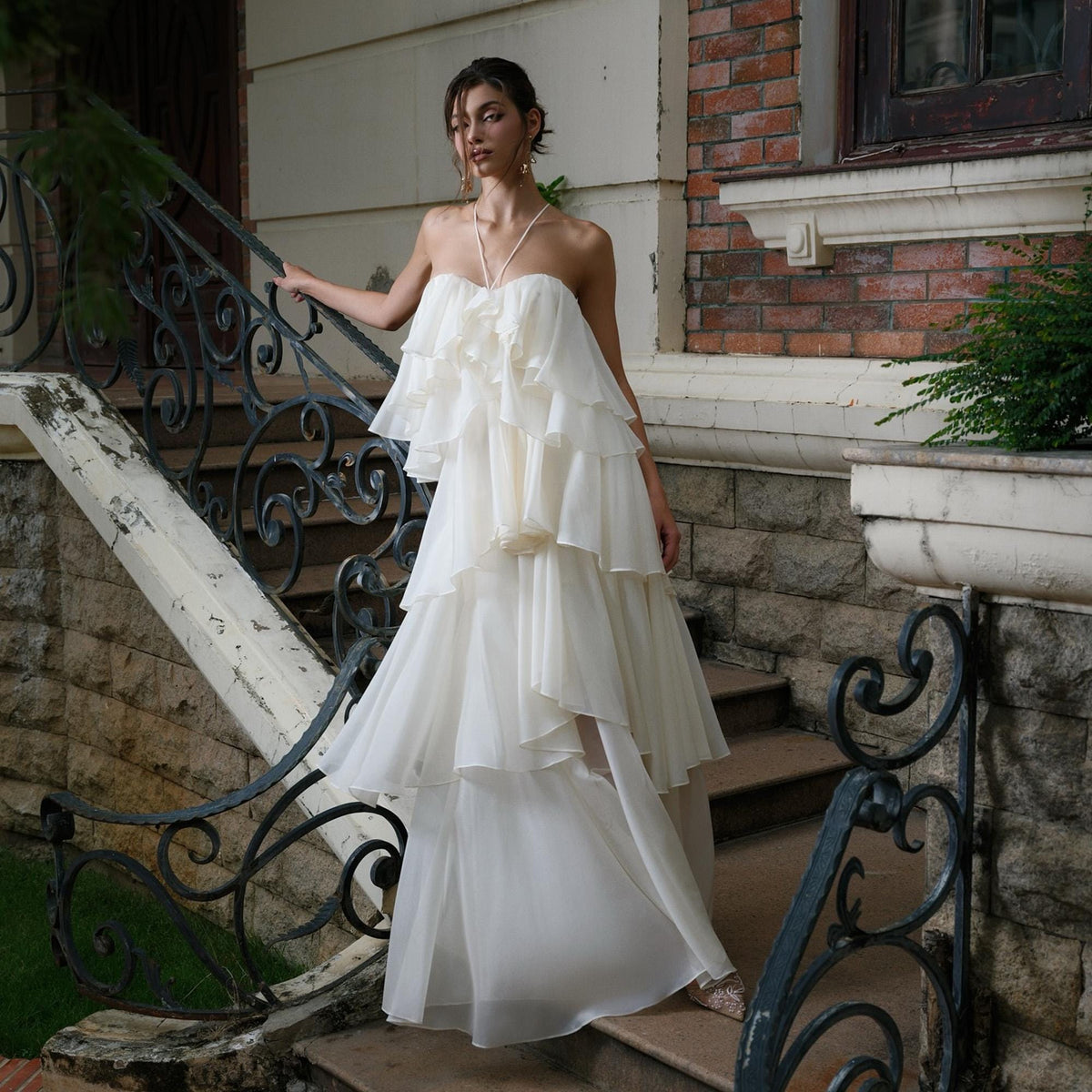 May include: An off-white, layered formal gown with a halter neckline. The dress features multiple tiers of ruffles and a flowing skirt. The model is standing on a stone staircase with a wrought iron railing, posing near a brick building.