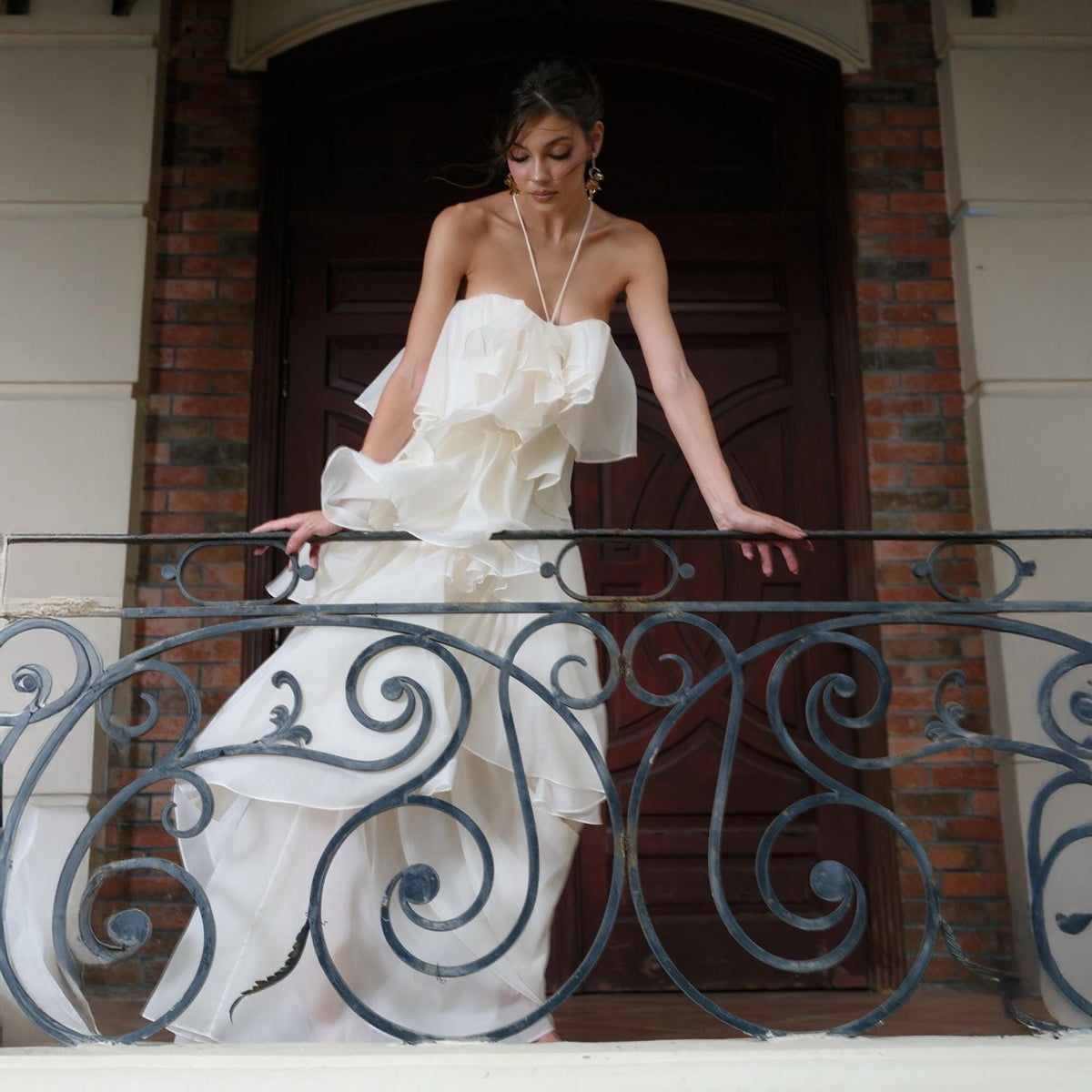 May include: A cream-colored, strapless gown with layered ruffles. The dress features a halter-style neckline and a flowing skirt. The model is standing on a balcony with a wrought iron railing, with a brick wall and wooden door in the background.