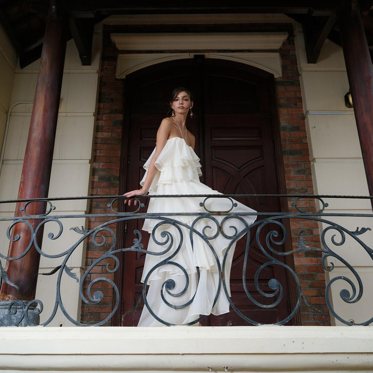 May include: A white, tiered, off-the-shoulder gown. The dress features multiple layers of flowing fabric and is worn on a balcony with ornate wrought iron railings. The background includes a dark wooden door and brickwork.