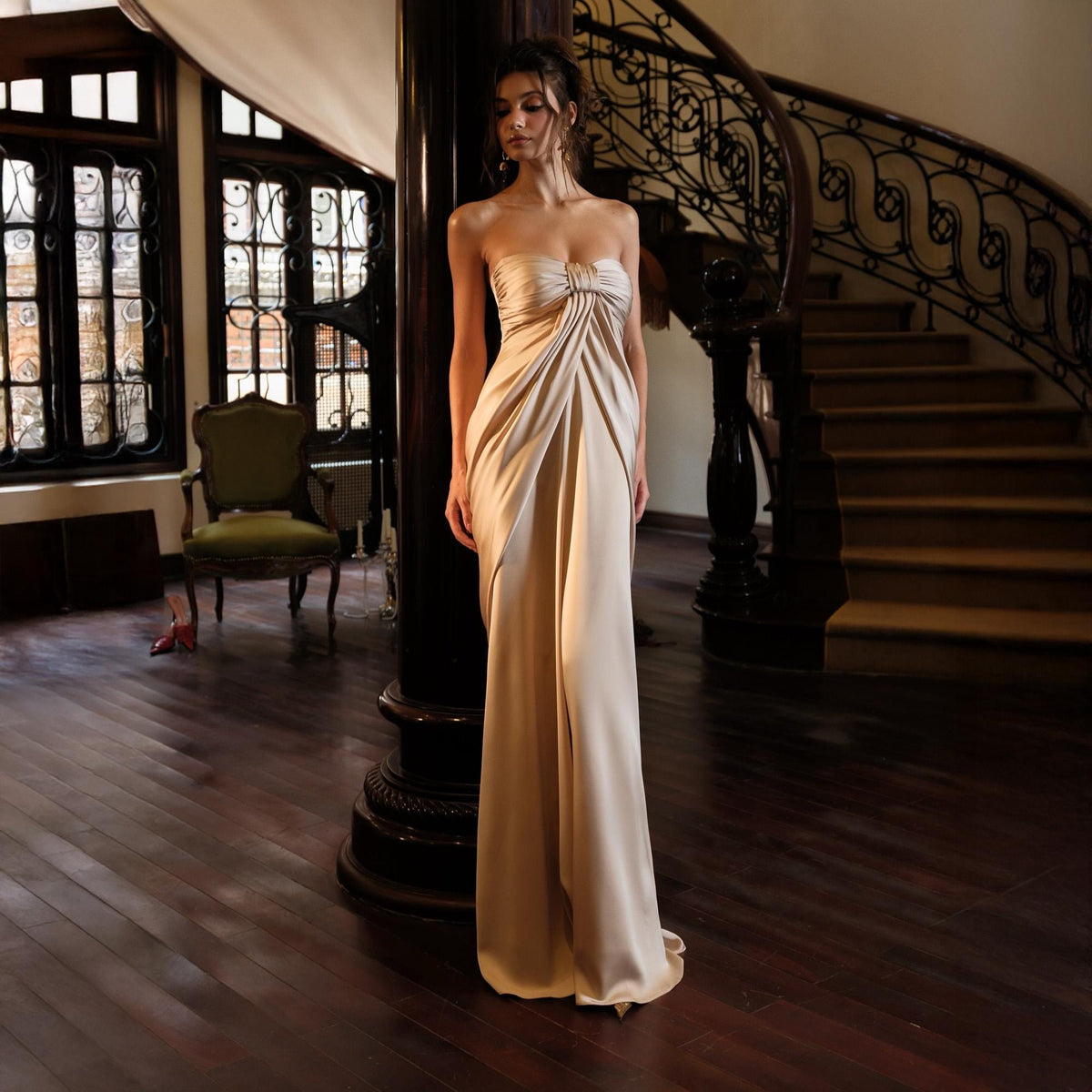 May include: A strapless, floor-length gown in a champagne color. The dress features a draped bodice and a flowing skirt. The model is standing in a room with a dark wood floor and a winding staircase in the background.