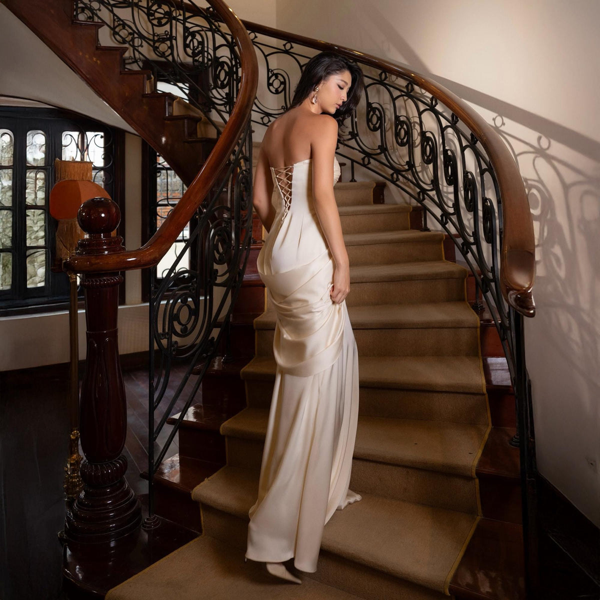 May include: A strapless ivory gown with a lace-up back descends a grand, curved staircase. The dress features a flowing skirt and is worn with matching heels. The staircase has a dark wood banister and ornate wrought iron detailing.