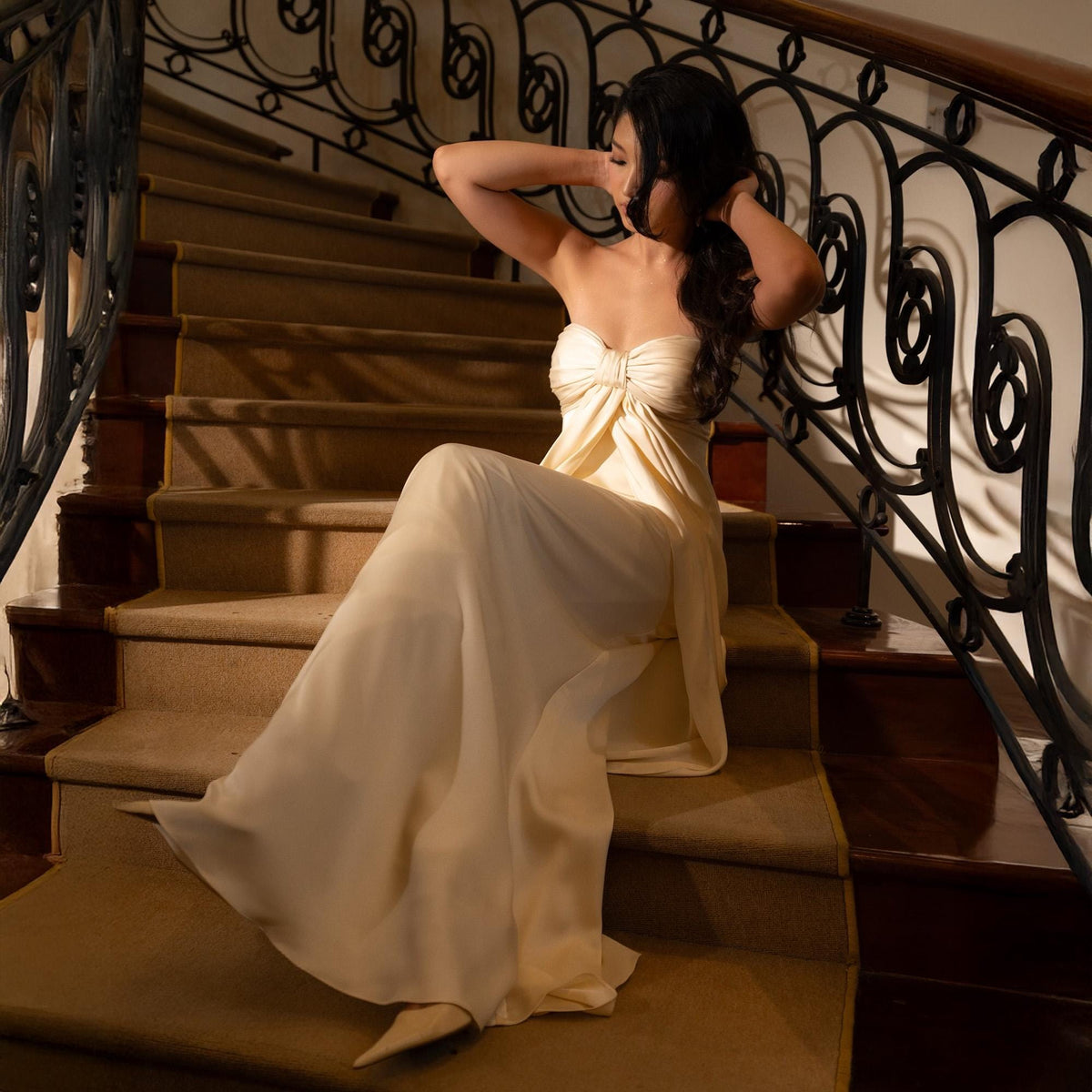 May include: A strapless, cream-colored gown with a flowing skirt. The dress features a bow detail at the bust and is worn on a staircase with a decorative wrought iron railing. The steps are covered in a neutral-toned carpet.