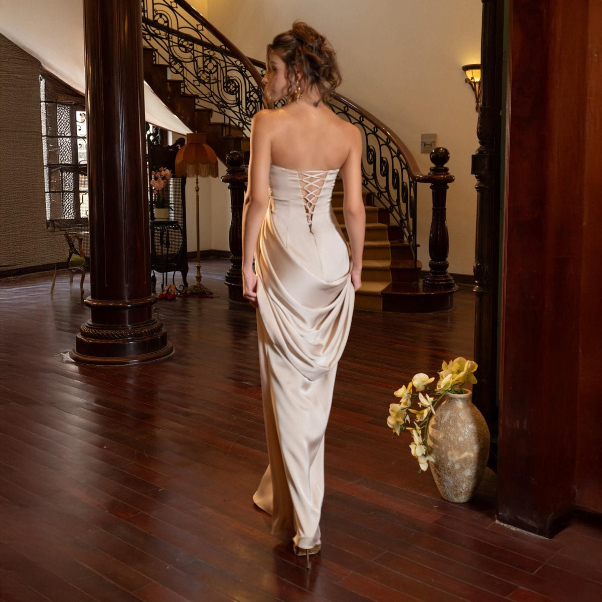 May include: A strapless, floor-length champagne-colored gown with a lace-up back. The dress features a draped skirt and is worn by a person walking away from the camera. The background includes a wooden floor, a staircase, and a decorative vase.