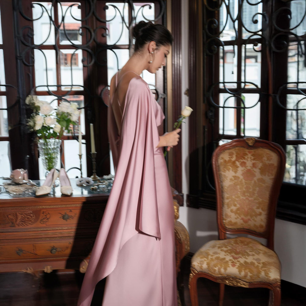 May include: A long, flowing, dusty rose-colored gown with cape sleeves. The dress features a low back and is worn with delicate jewelry. A white rose is held in the hand. The setting is a room with antique furniture and large windows.