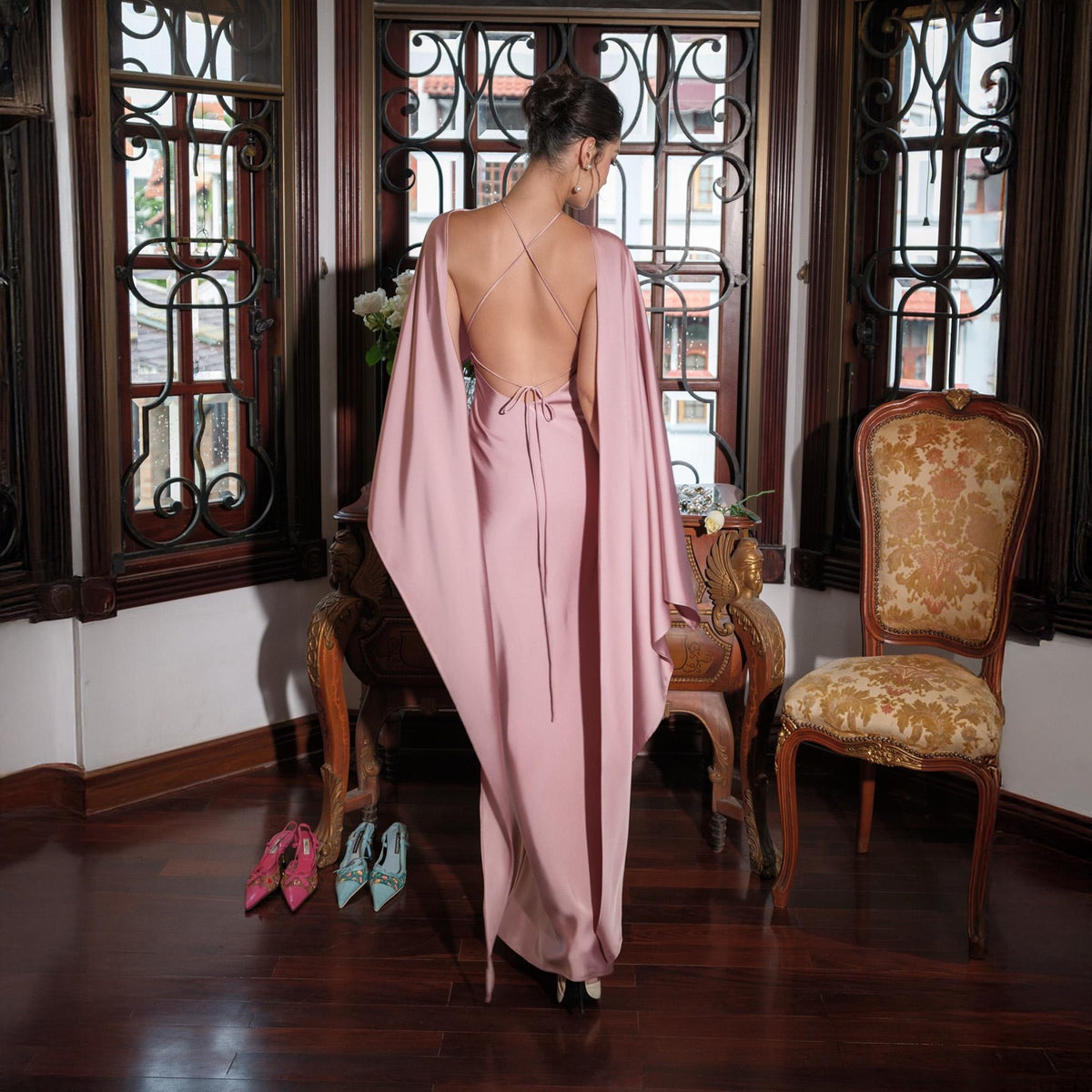 May include: A dusty rose-colored formal gown with a flowing cape-like design. The dress features a low back with delicate strap detailing. The model is standing in a room with ornate windows and antique furniture. Two pairs of high heels are on the floor.